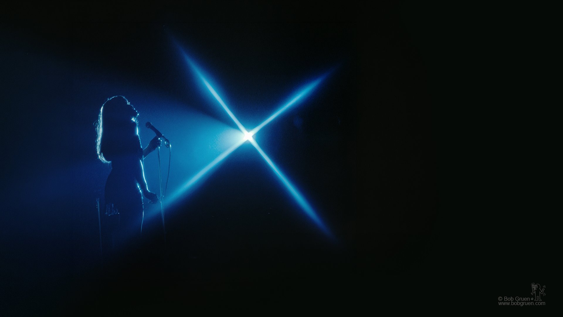 Tina Turner in silhouette against a cross of blue stage light — photograph by Bob Gruen